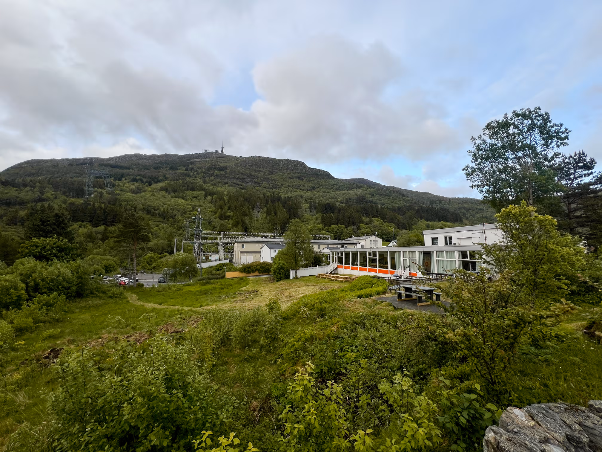 Modern buildings by a green landscape with mountains in the background under a cloudy sky.