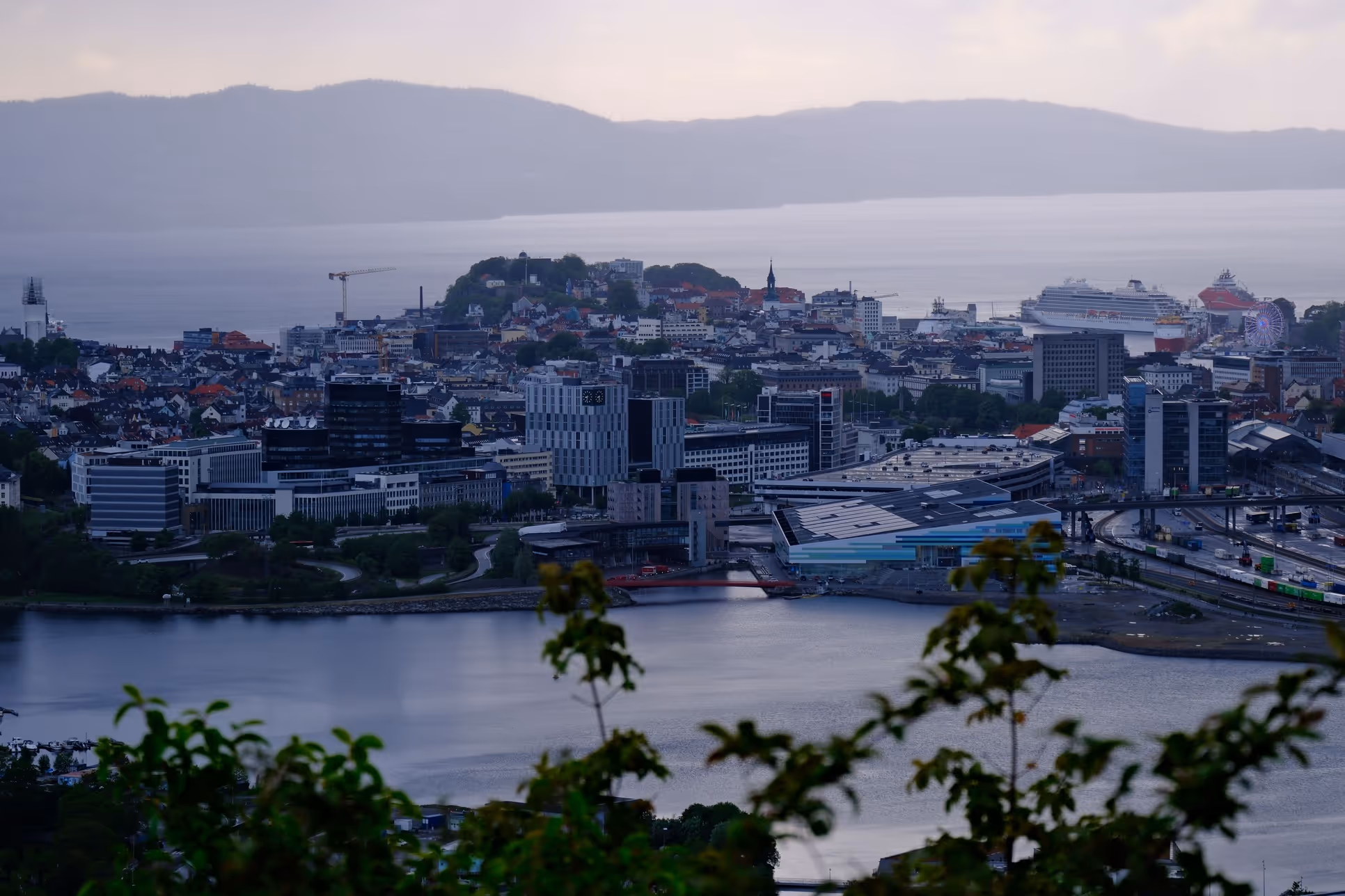 View of a city with modern and historic buildings by the water, cruise ships in the harbor, and green mountains in the background.