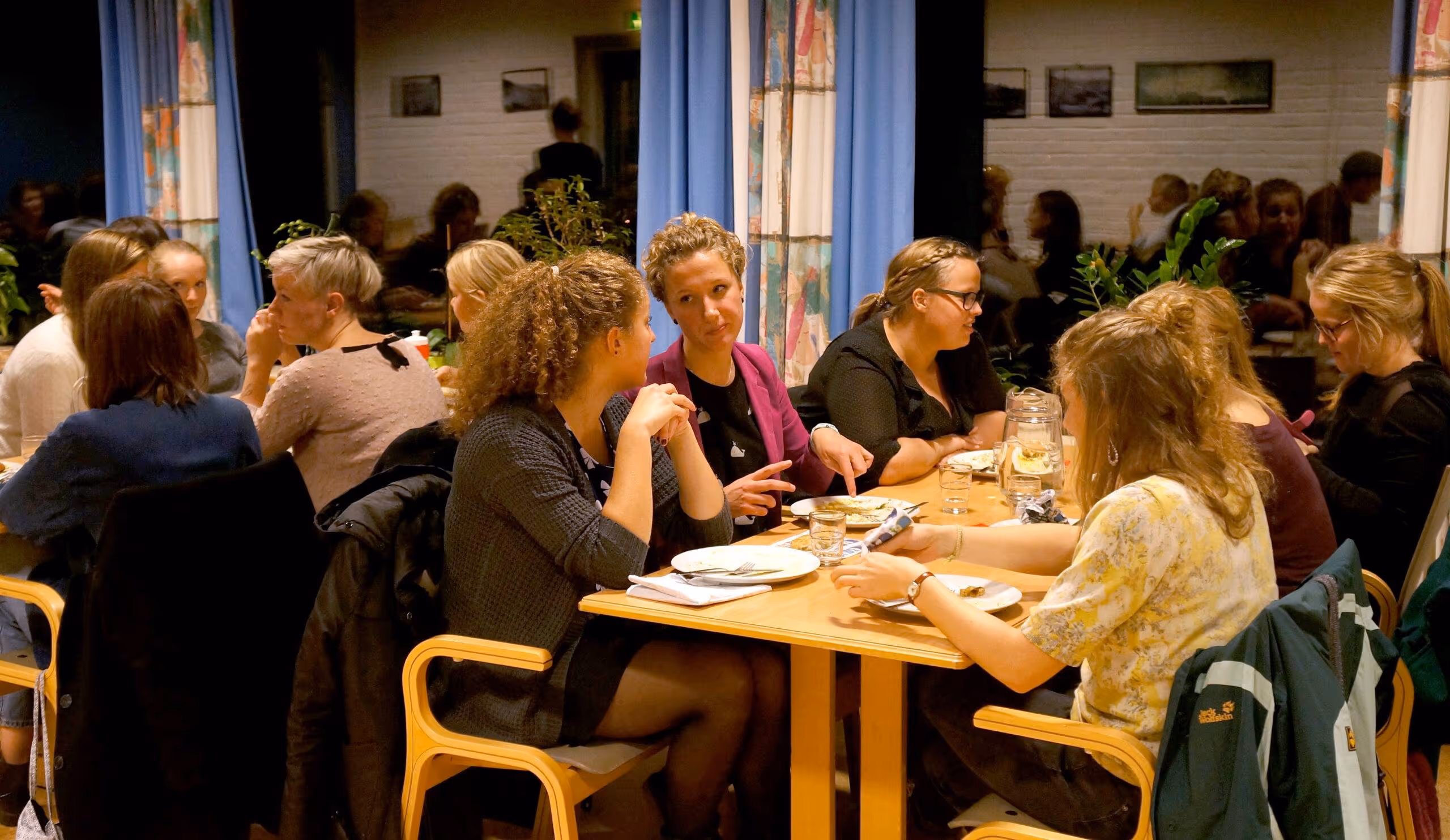 Gruppe unge voksne som sitter rundt et bord og snakker under et måltid innendørs.