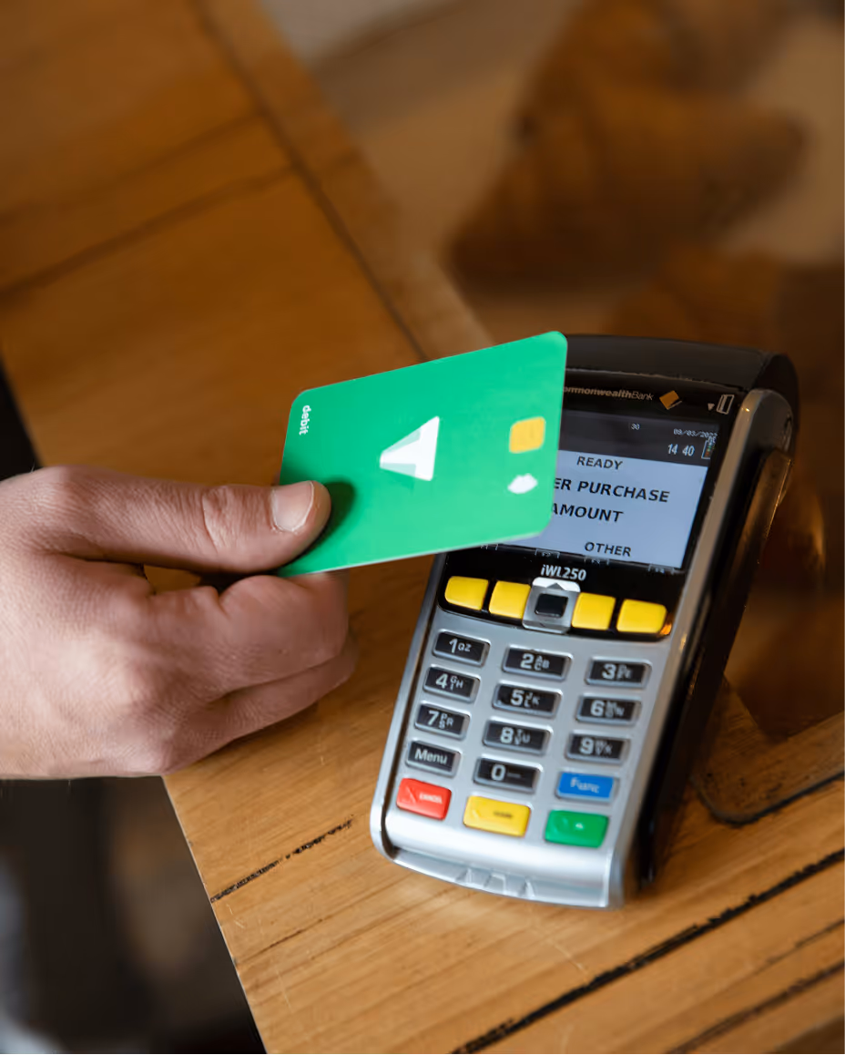 Hand holding green debit card near a payment terminal on a wooden counter for contactless payment.