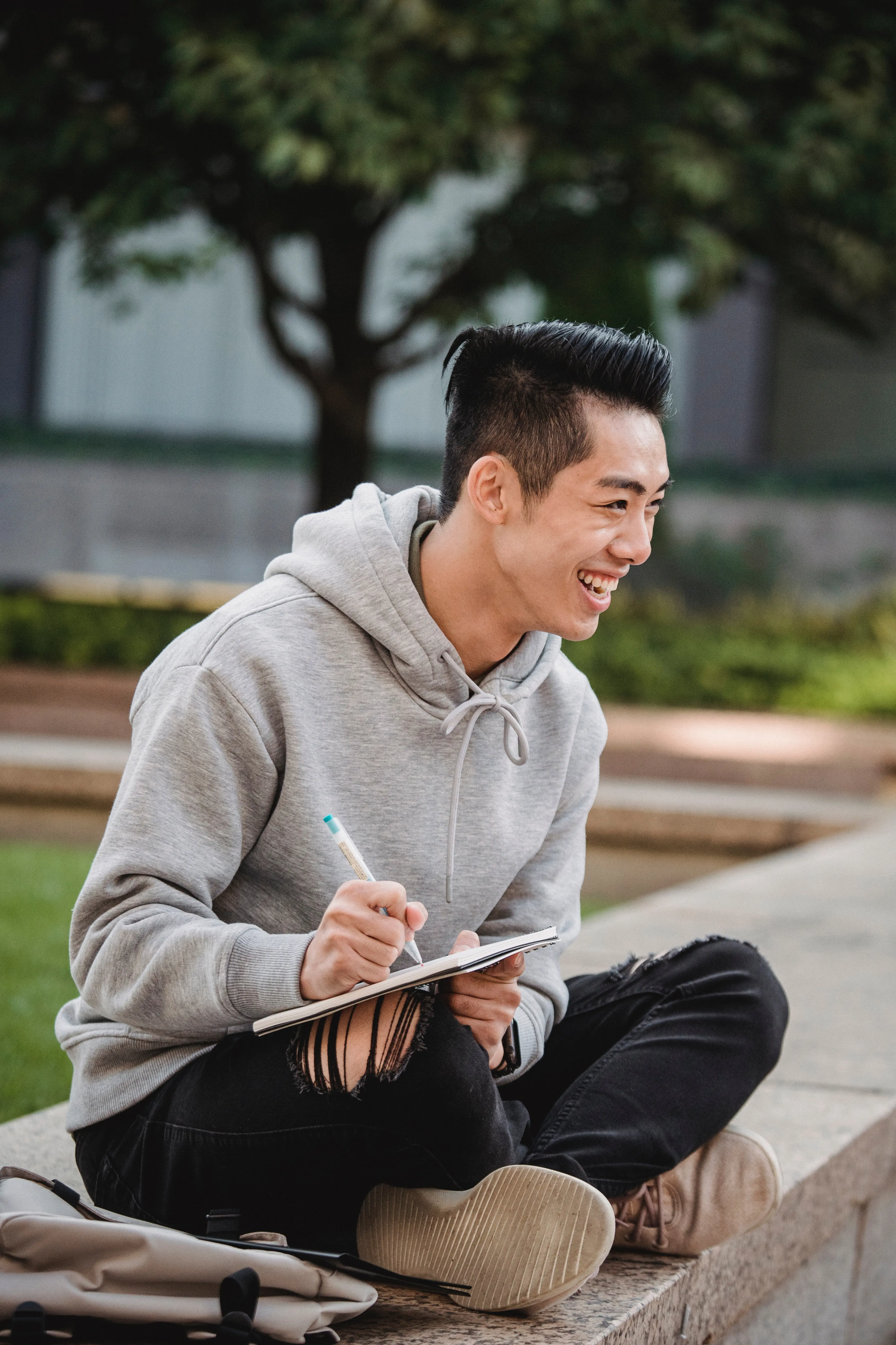 Young man in a gray hoodie and ripped black jeans sitting cross-legged on a stone bench, smiling while writing in a notebook outdoors.