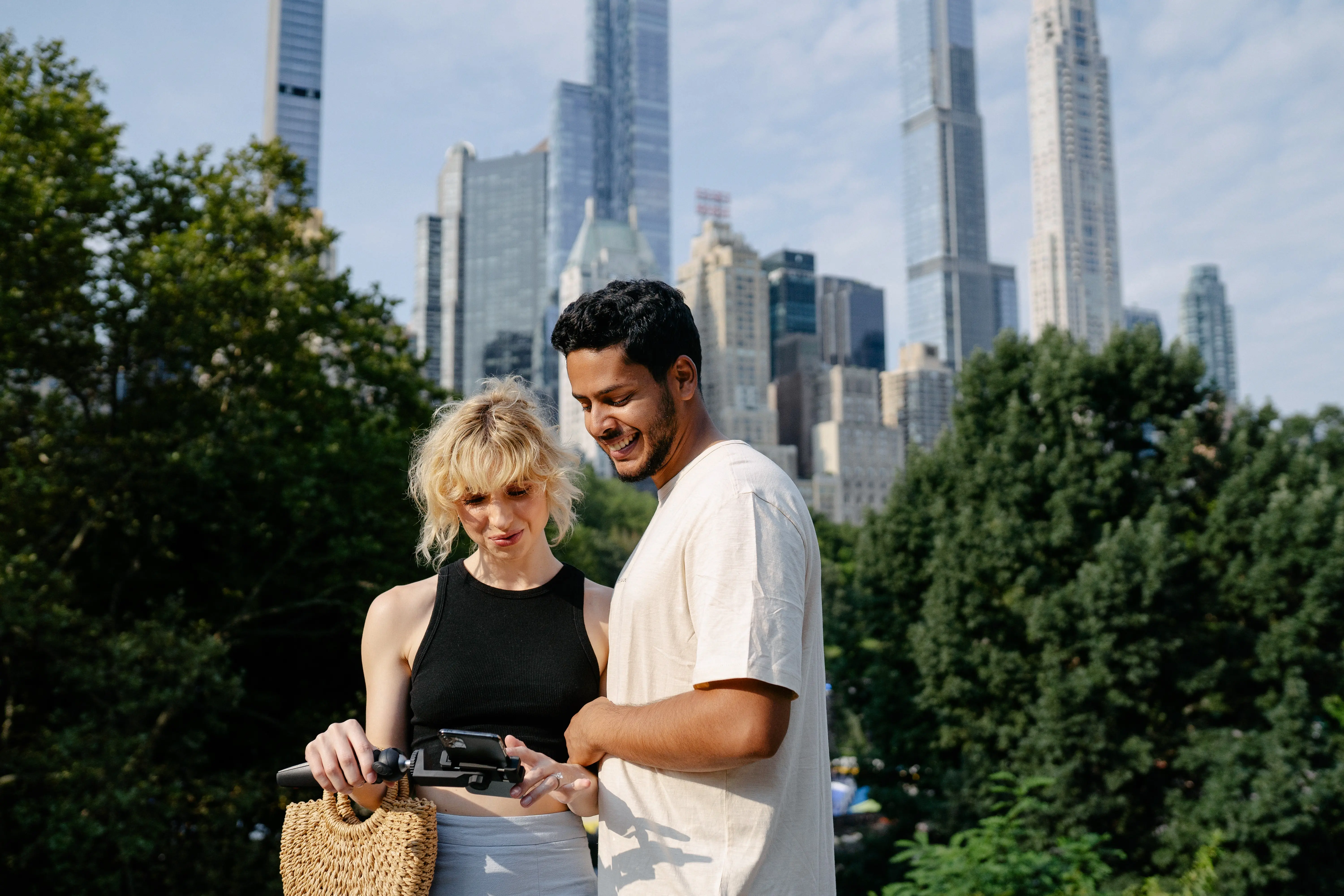 Smiling couple using a smartphone together outdoors with city skyscrapers and trees in the background.