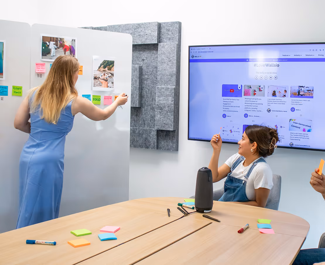 A woman in a blue dress writes on a whiteboard covered with pictures and colorful sticky notes, while another woman in overalls sits at a table holding a small note, with a large screen displaying a social media dashboard on the wall.