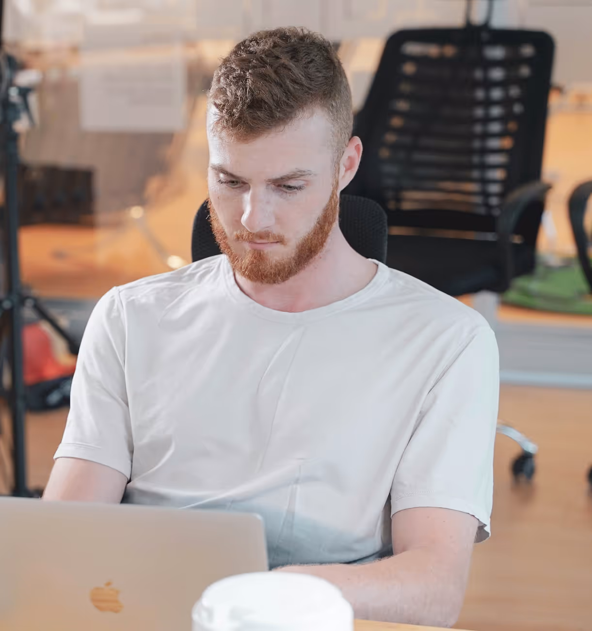 Young man with red beard and curly hair focused on his laptop in a modern office setting.