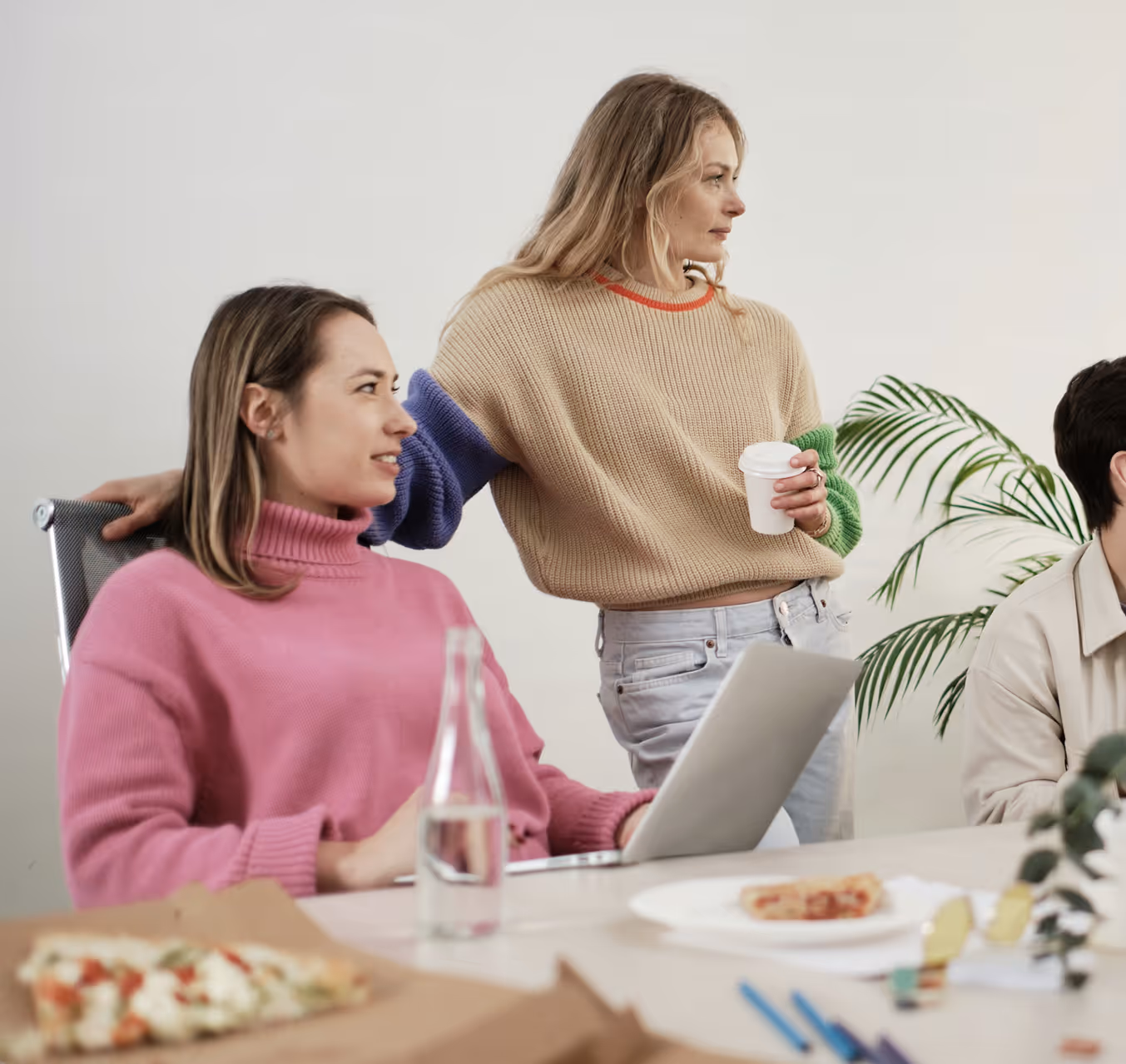 Two women in casual sweaters, one seated using a laptop and the other standing holding a coffee cup, with a pizza and water bottle on the table.