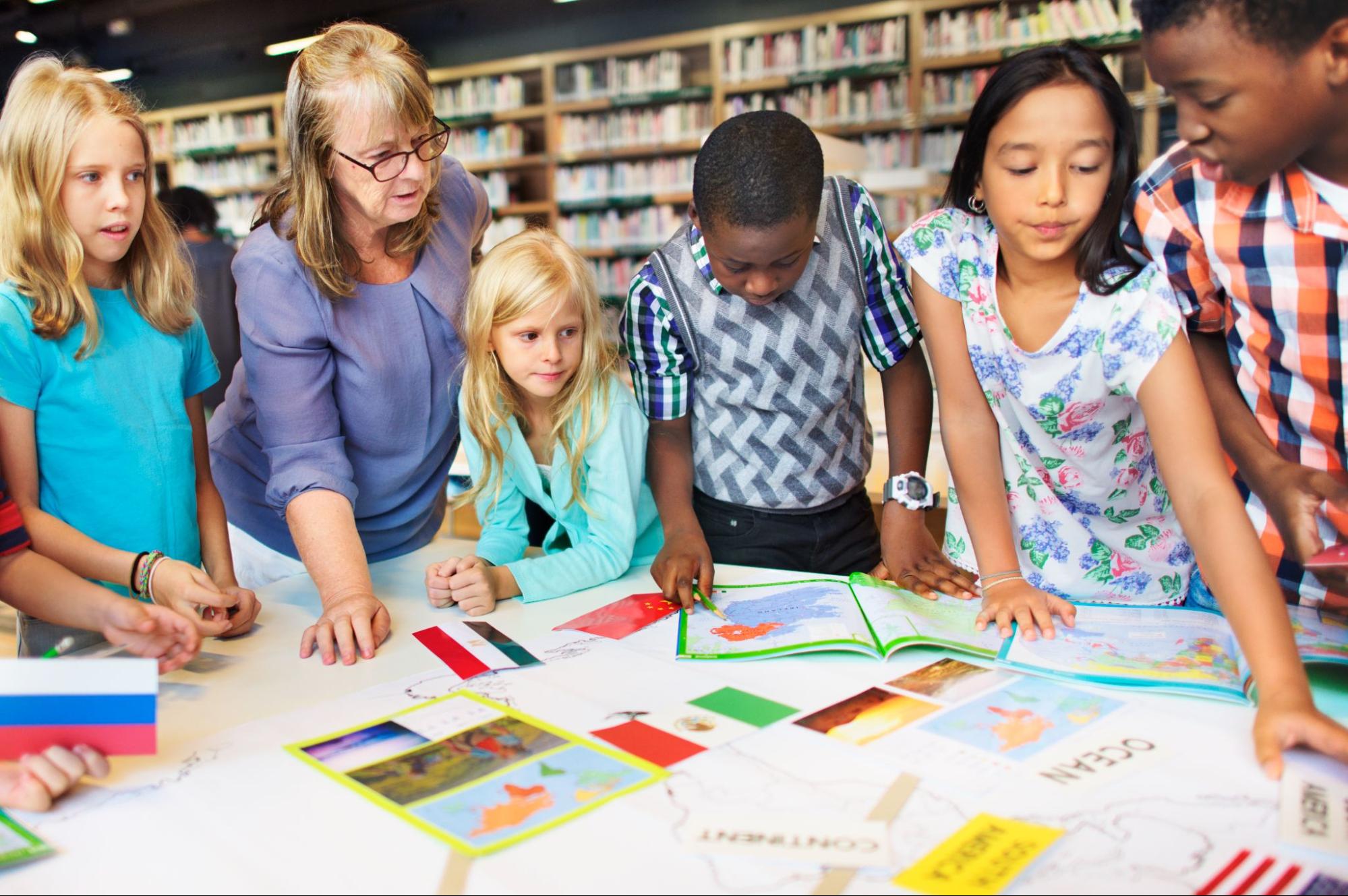 A group of children and a teacher look at maps and flags on a table.