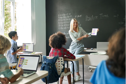 Smiling teacher sitting on a desk while holding iPad and giving a math lesson to students