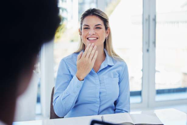 Deaf employee saying Thank you to a colleague in sign language. She is wearing a blue blouse and sitting at a white desk with a notebook open in front of her.