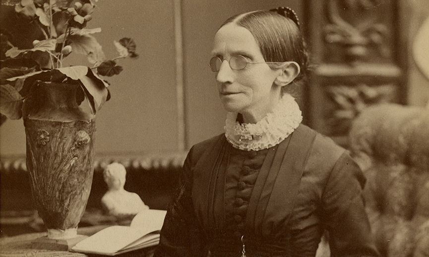 vintage portrait of a woman with glasses in Victorian dress standing beside a vase and open book