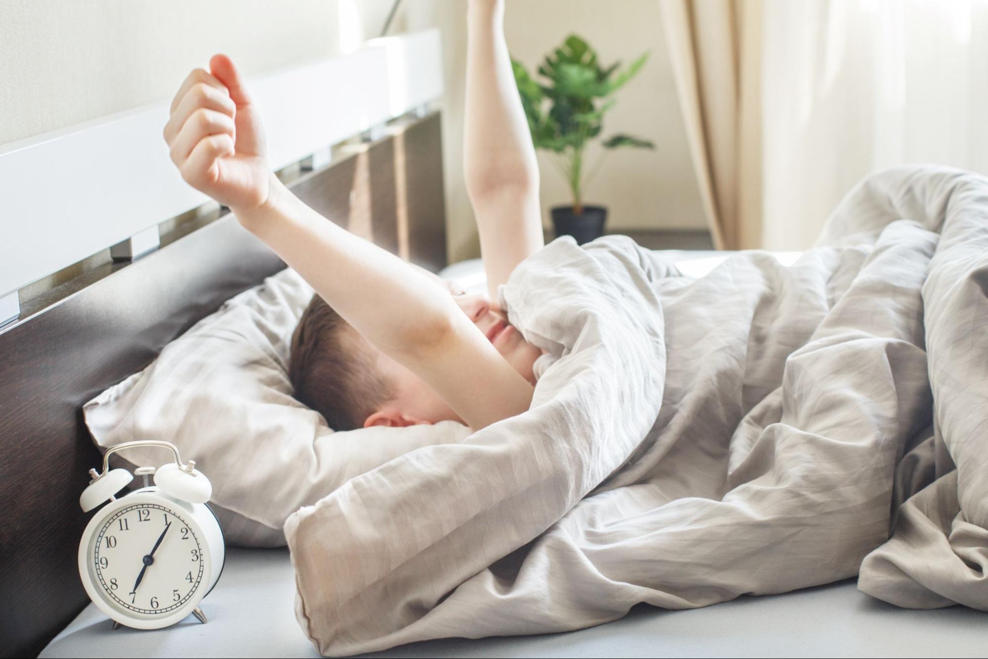 A little boy waking up in bed next to an alarm clock.