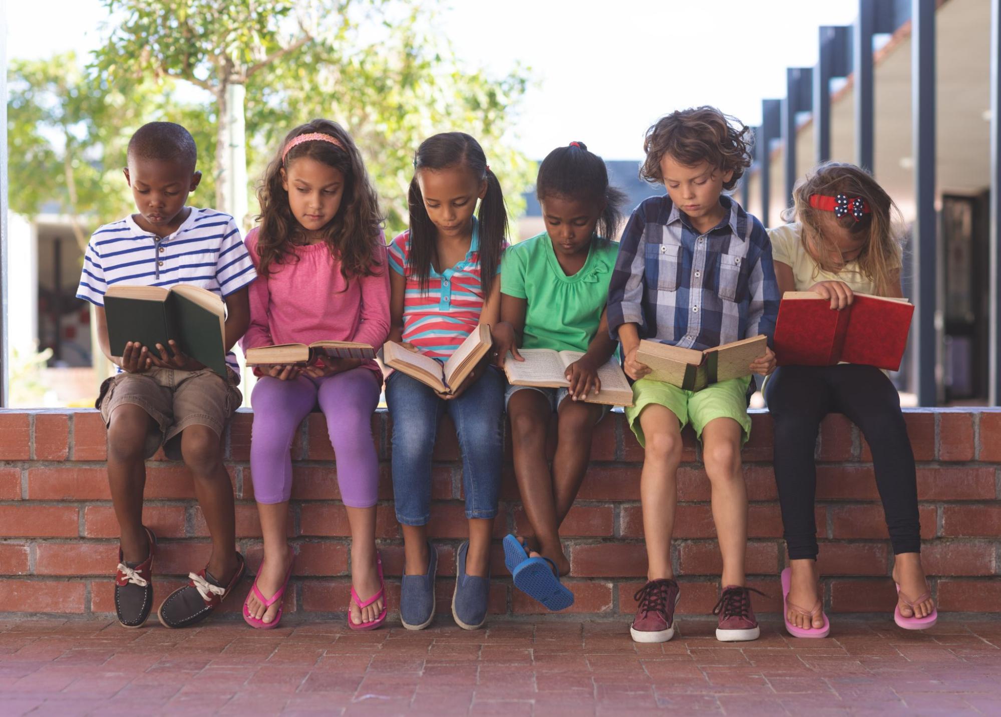 A group of young children sitting on a brick wall and reading books.