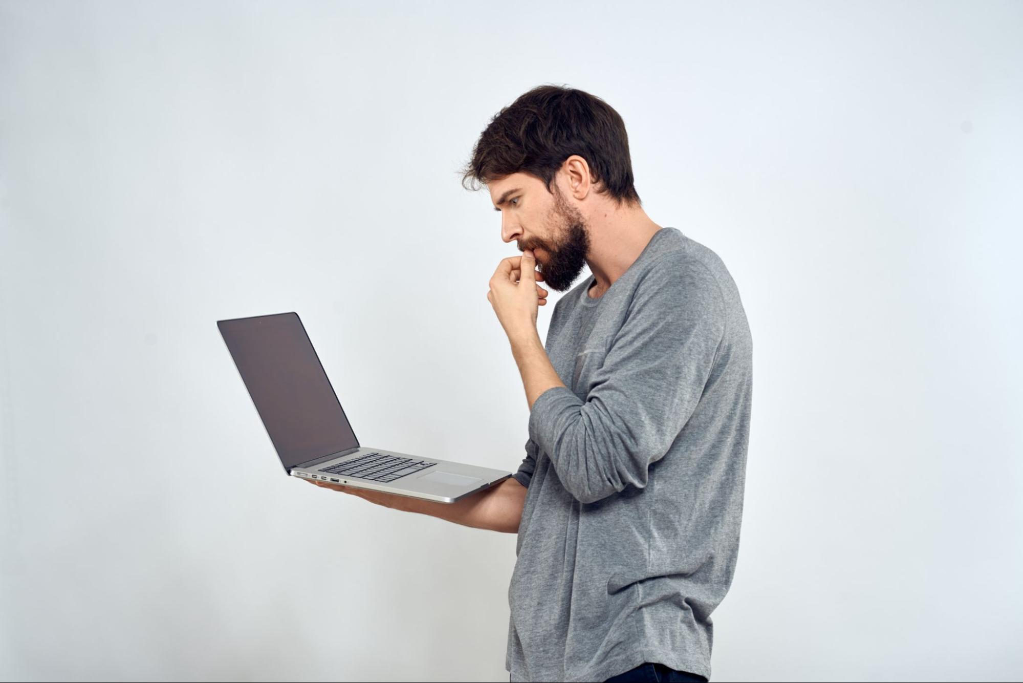 A bearded white man with dark hair and a gray shirt is holding a laptop computer and thinking.