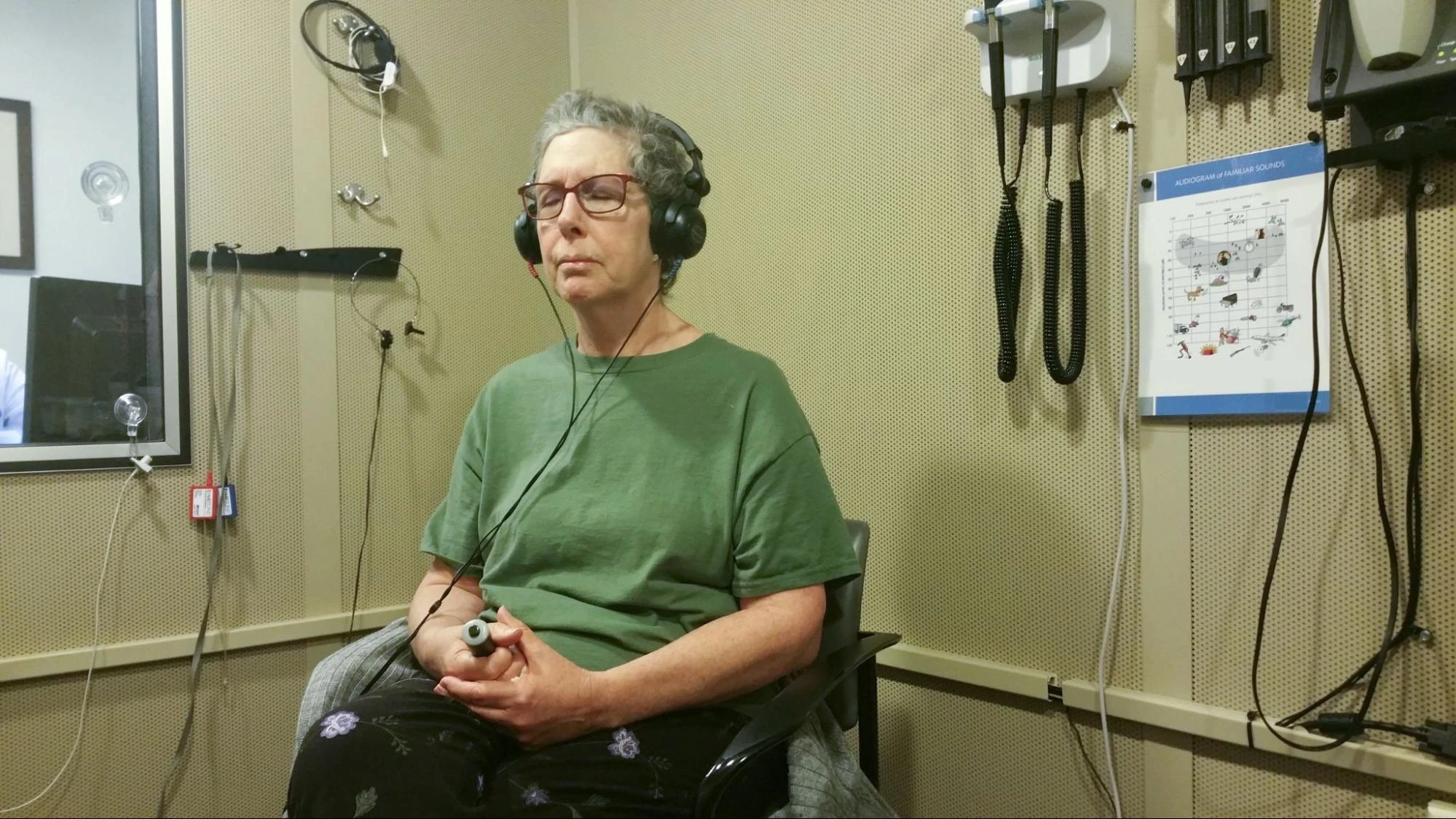 Patient wearing headphones and holding a response button during an audiogram hearing test inside a soundproof audiology examination room.