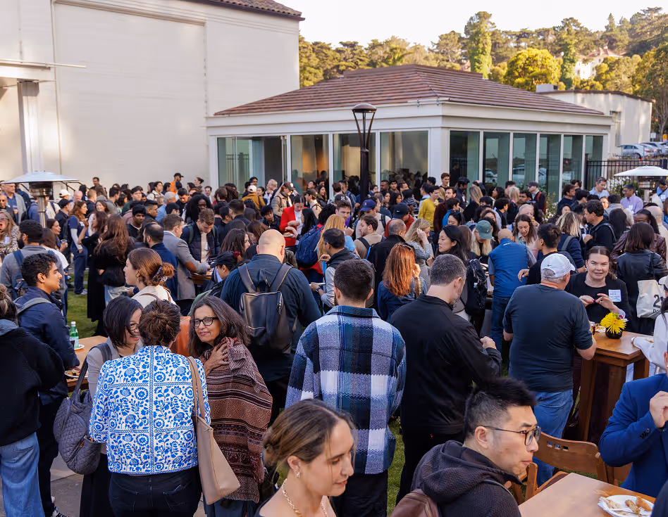 Large diverse crowd of people socializing outdoors near a building with glass walls during daytime.