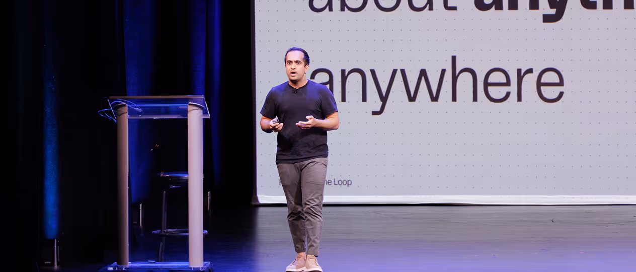 Man in black shirt and gray pants speaking on stage with a large screen behind showing the words 'about anything anywhere'.