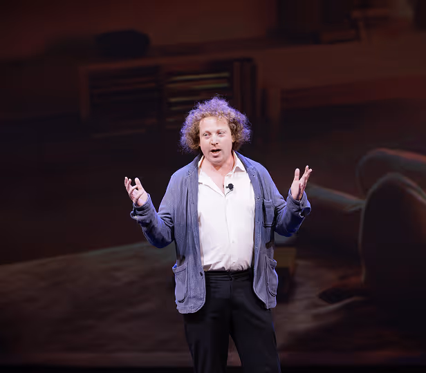 Man with curly hair speaking on stage with expressive hand gestures, wearing a blue cardigan and white shirt.