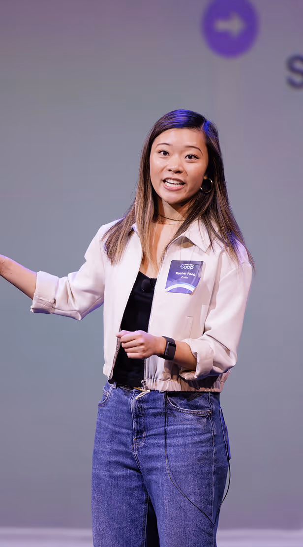 Woman with long hair wearing a light jacket and jeans speaking on stage with a microphone headset and a badge reading 'Rachel Pong'.