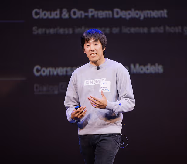 Man wearing a gray sweatshirt speaking on stage in front of a presentation screen with text about cloud and on-prem deployment.