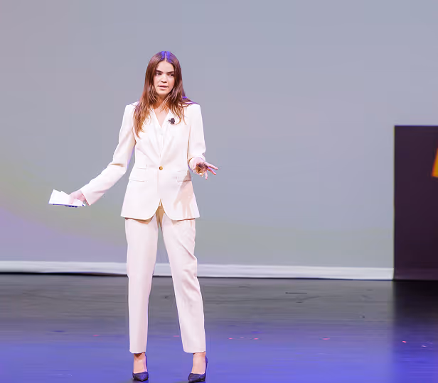 Woman in a white suit speaking on stage with a microphone, holding papers in one hand.