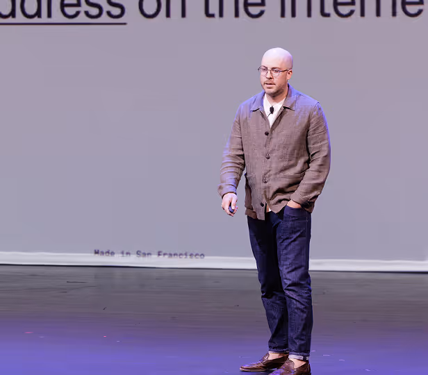 Man with glasses and shaved head wearing a brown jacket and blue jeans speaking on stage with a presentation screen behind him.