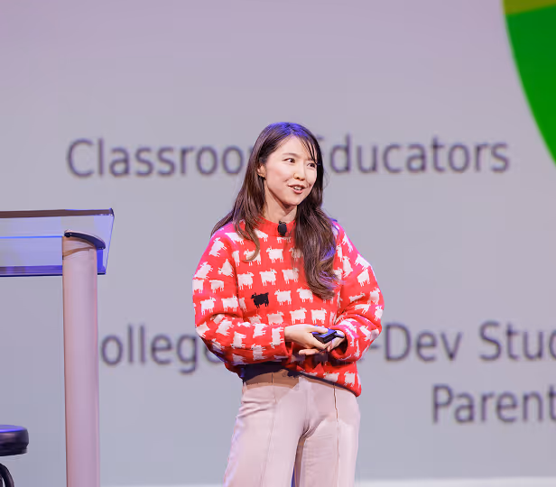 Woman wearing a red sweater with white and black sheep patterns speaking on stage in front of a presentation screen.