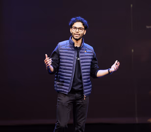 Man with glasses wearing a blue puffer vest and black clothes speaks with open hands against a dark background.