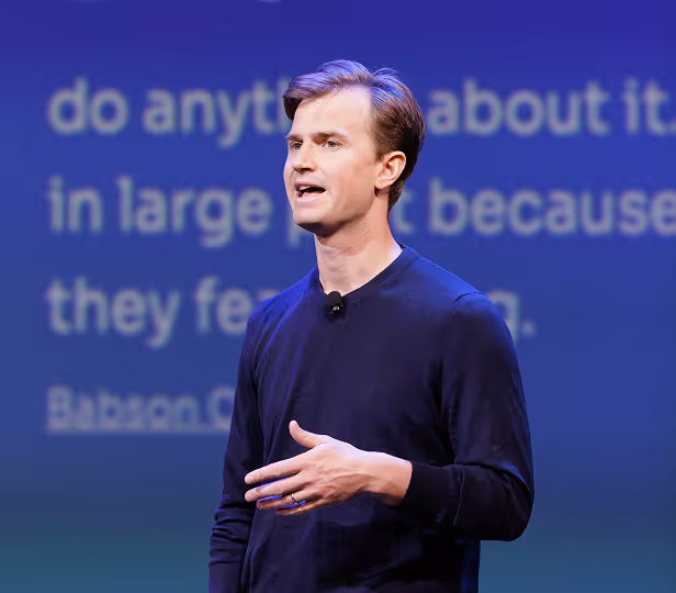 Man in a dark long-sleeve shirt speaking on stage with a blurred blue background displaying partially visible text.