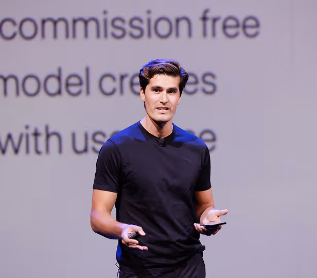 Man in a black t-shirt speaking on stage with a blurred presentation screen behind him.