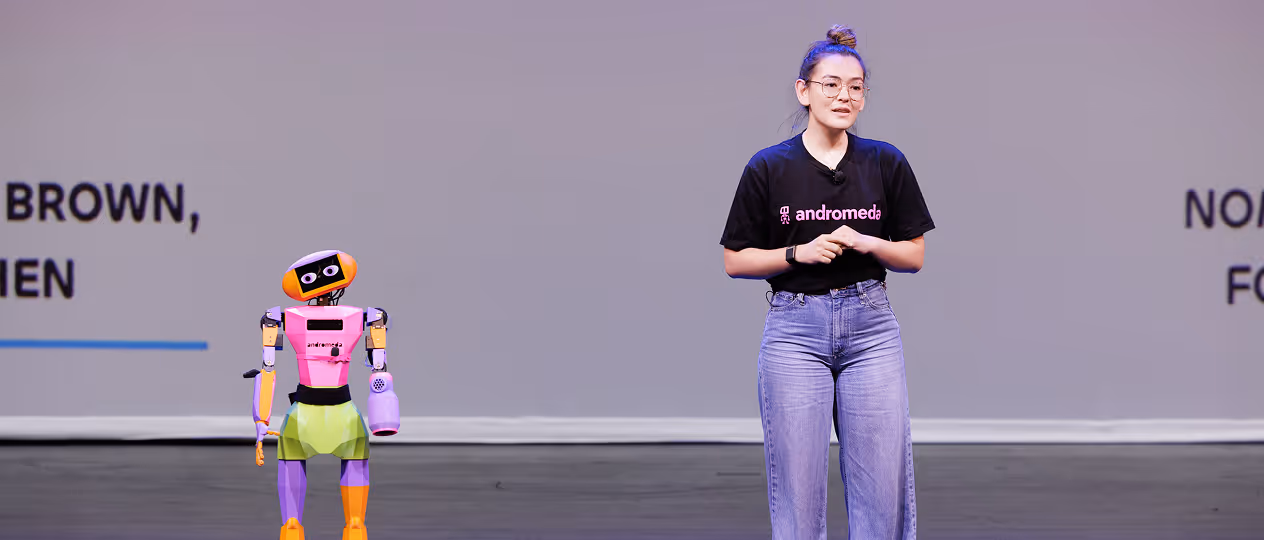 Woman with glasses and hair in a bun speaking on stage next to a small colorful humanoid robot.