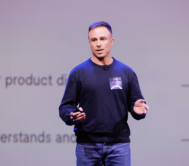 Man wearing a dark sweatshirt and jeans speaking on stage with hands gesturing in front of a blurred presentation screen.