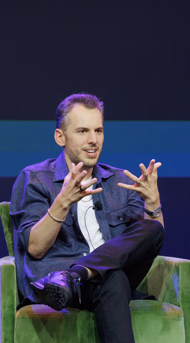 Man sitting cross-legged on a green chair speaking and gesturing with hands.