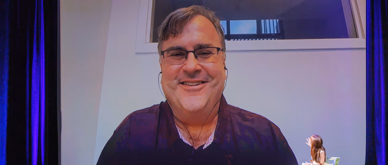 Man with glasses and a headset smiling on a large screen, with a woman sitting and gesturing at the bottom right corner.