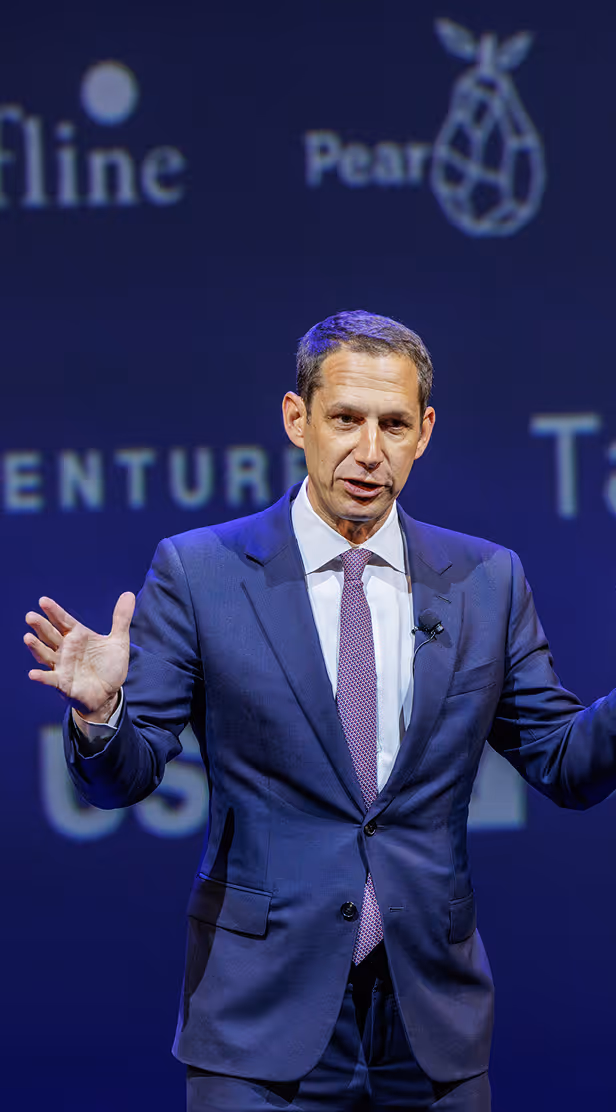 Man in a navy suit and purple tie speaking on stage with a dark blue background and projected logos.