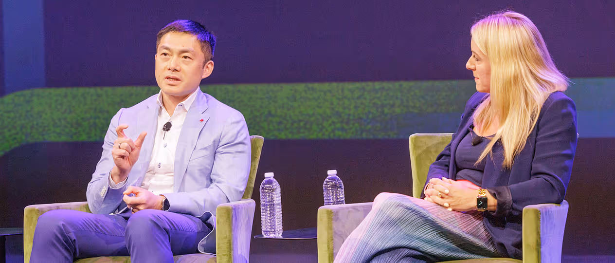 Two people sitting on green chairs on a stage, one man in a light blue blazer speaking and gesturing with his hand, a woman with blonde hair listening attentively.