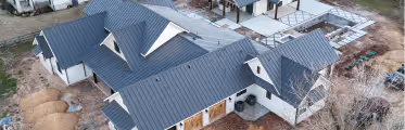Aerial view of a modern residential house with dark metal roofing and white walls, surrounded by bare trees and construction materials.