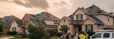 A fallen tree resting on the roof of a suburban house while several people stand nearby assessing the damage at sunset.