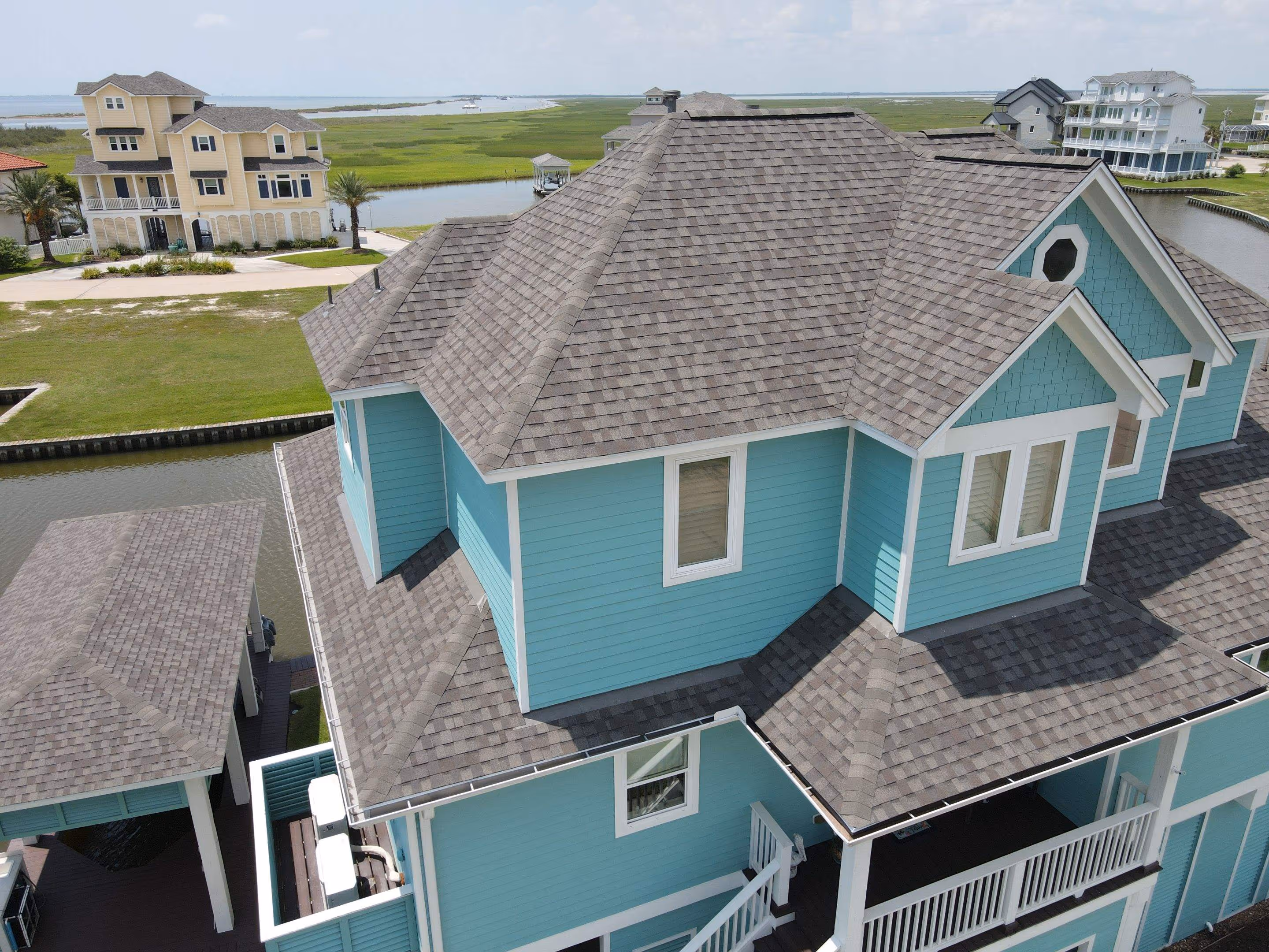 Aerial view of a blue coastal house with gray shingle roof near water and neighboring houses in the background.