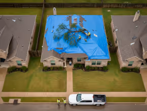 Aerial view of a house with a blue tarp covering the roof and workers cleaning up tree debris.