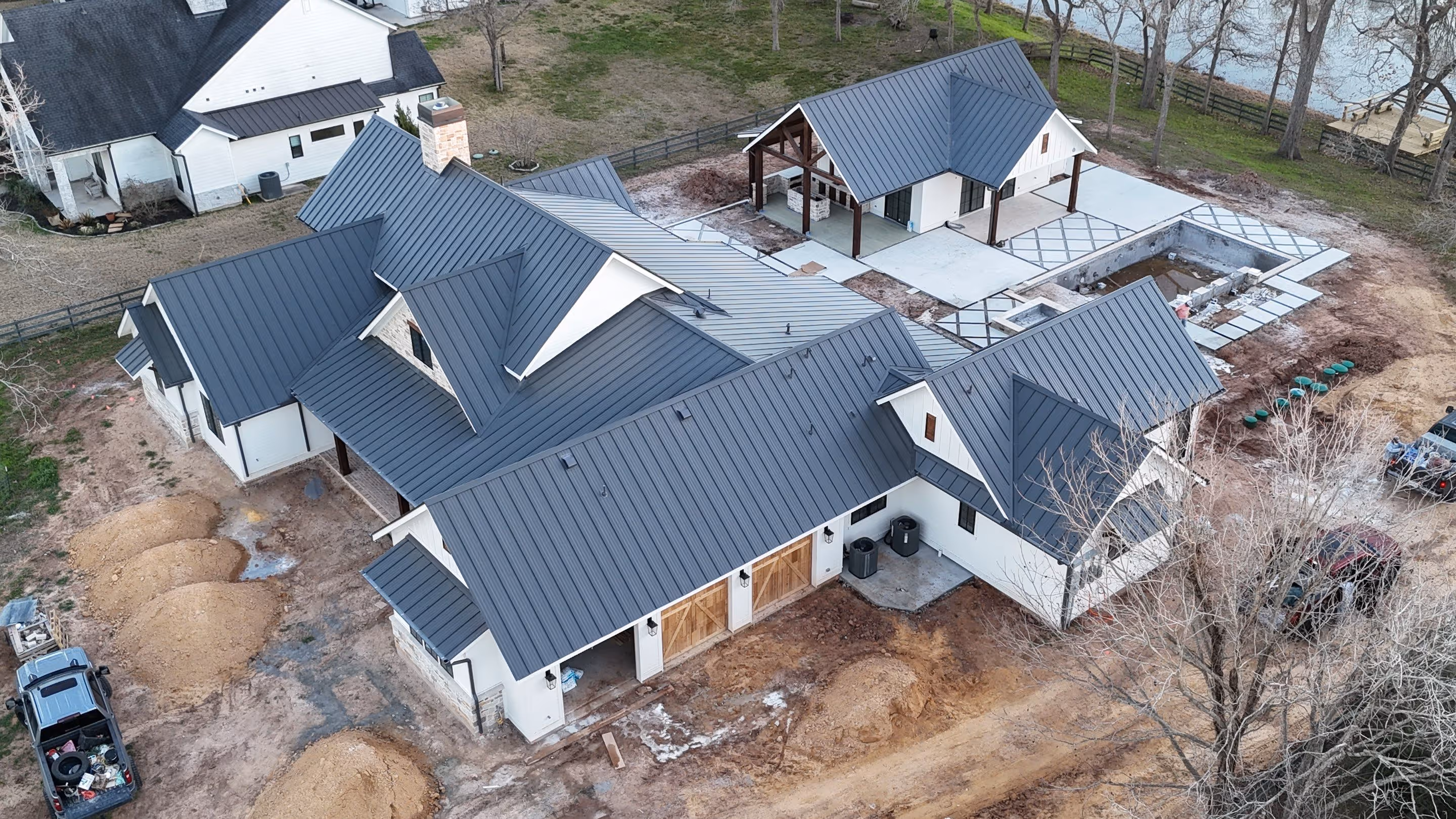 Aerial view of a large house under construction with metal roofing, an unfinished pool, and surrounding dirt and construction materials.