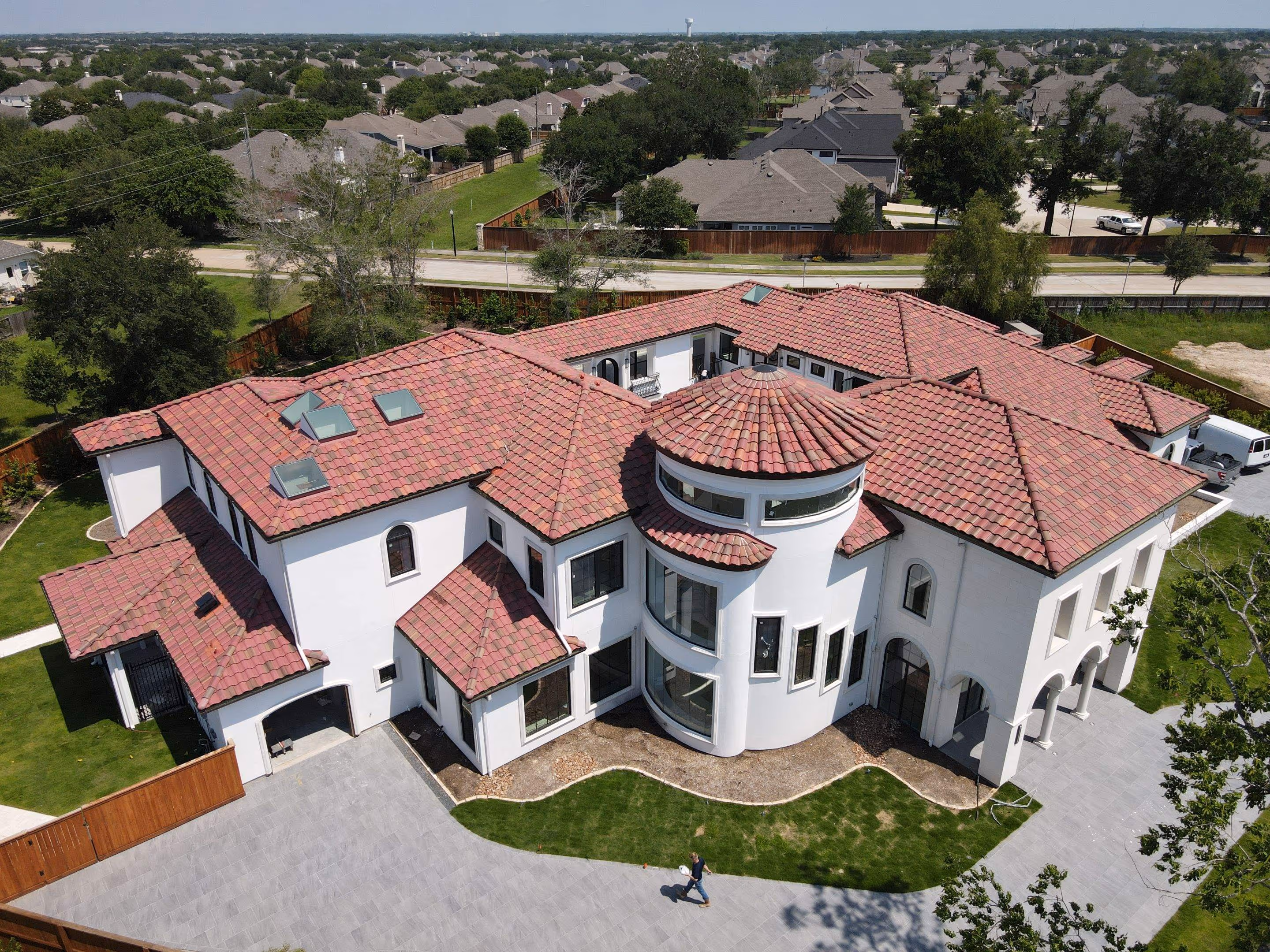Aerial view of a large white luxury house with red tile roofing, curved tower windows, and a paved driveway.