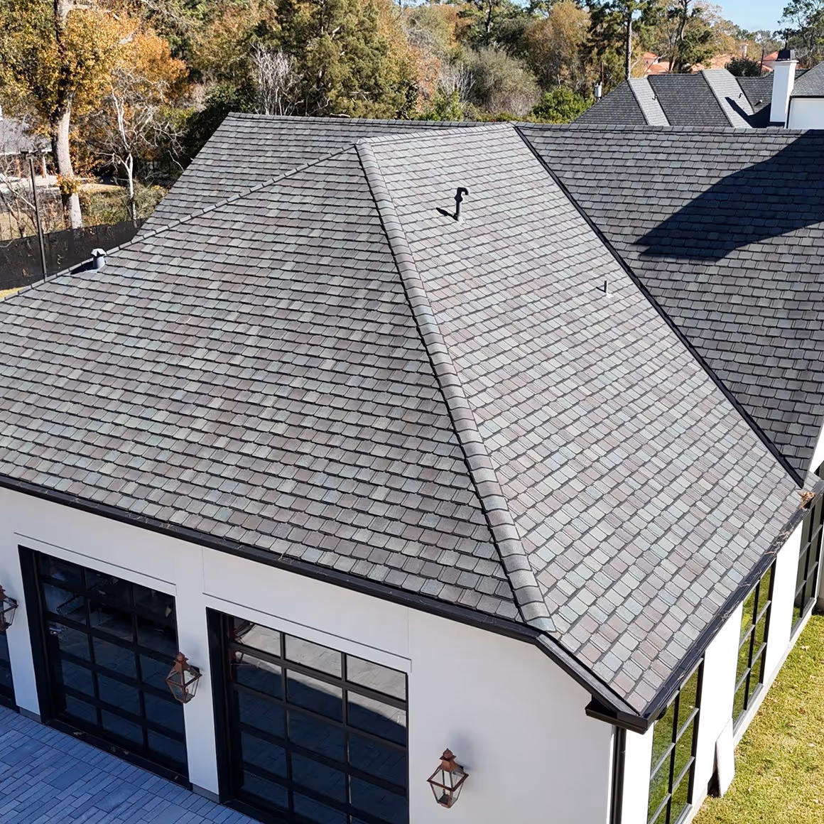 Aerial view of a house with a gray shingled hip roof, white exterior walls, and black framed garage doors and windows.