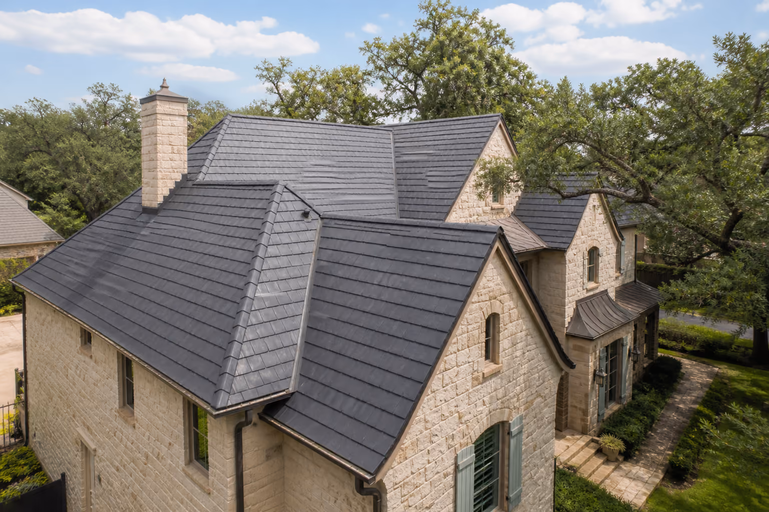 Stone house with dark synthetic roofing tiles and green window shutters surrounded by trees and grass.