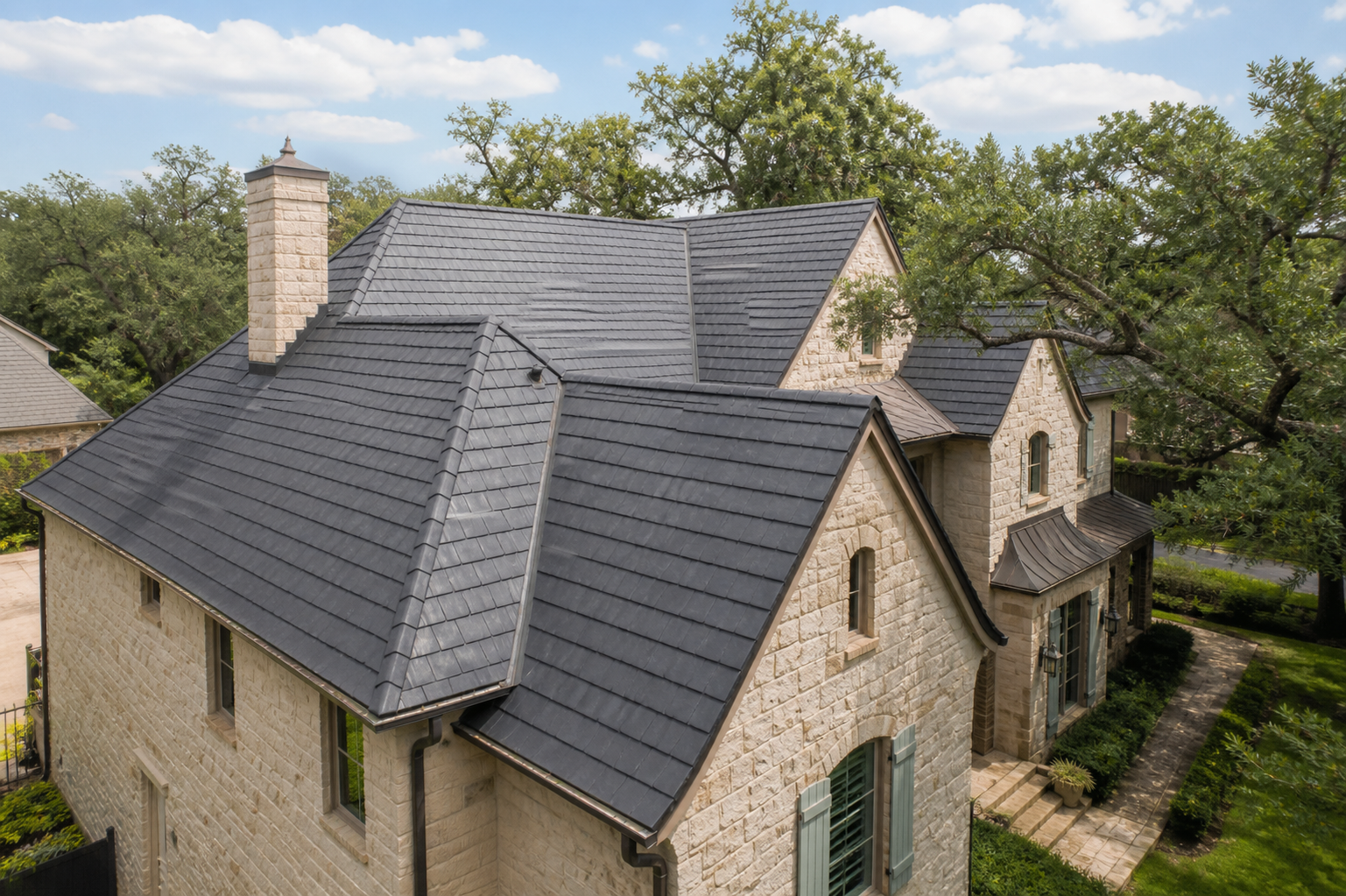 Stone house with dark synthetic roofing tiles and green window shutters surrounded by trees and grass.