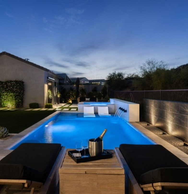 Modern pool with water feature and lounge chairs at dusk beside a desert hillside by Kevens Landscape