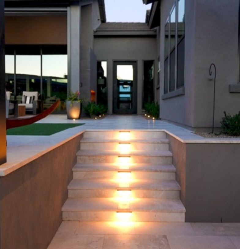Lit travertine steps leading to a raised patio with artificial turf by Kevens Landscape