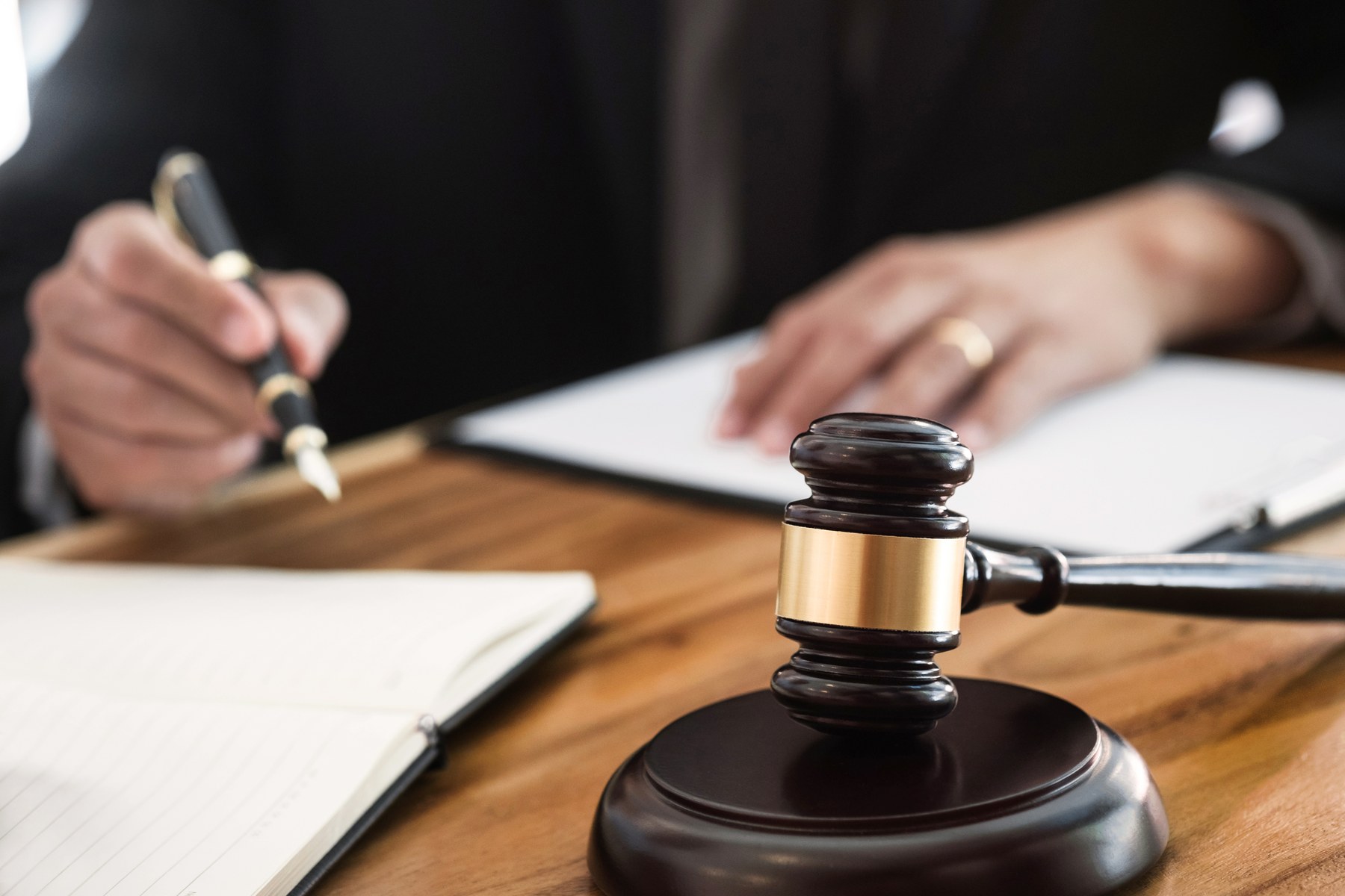 Gavel on a wooden desk with a person writing on paper, pen in hand. Formal setting suggests legal or judicial environment.