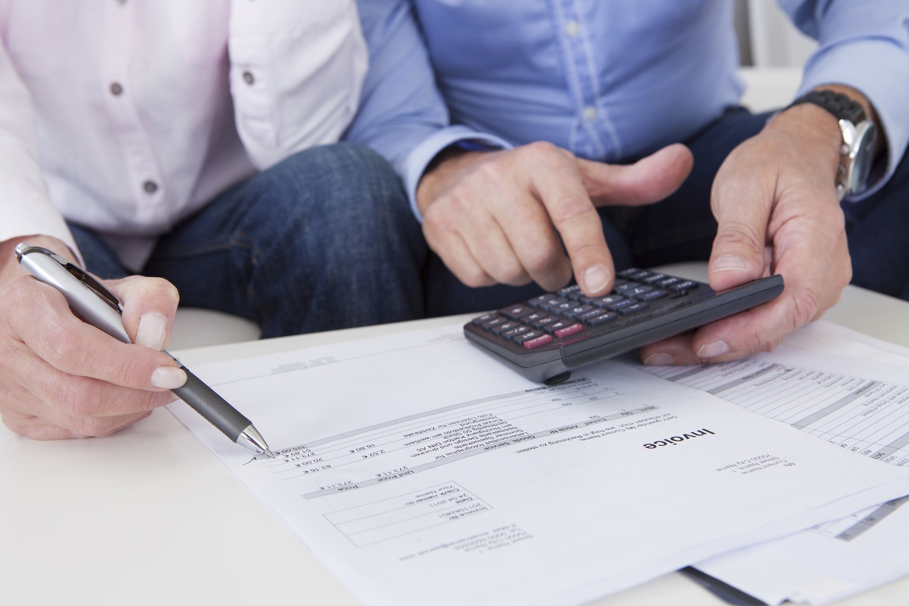 Two people calculating on an invoice with a pen and calculator in a bright office. Focus on hands and document details.