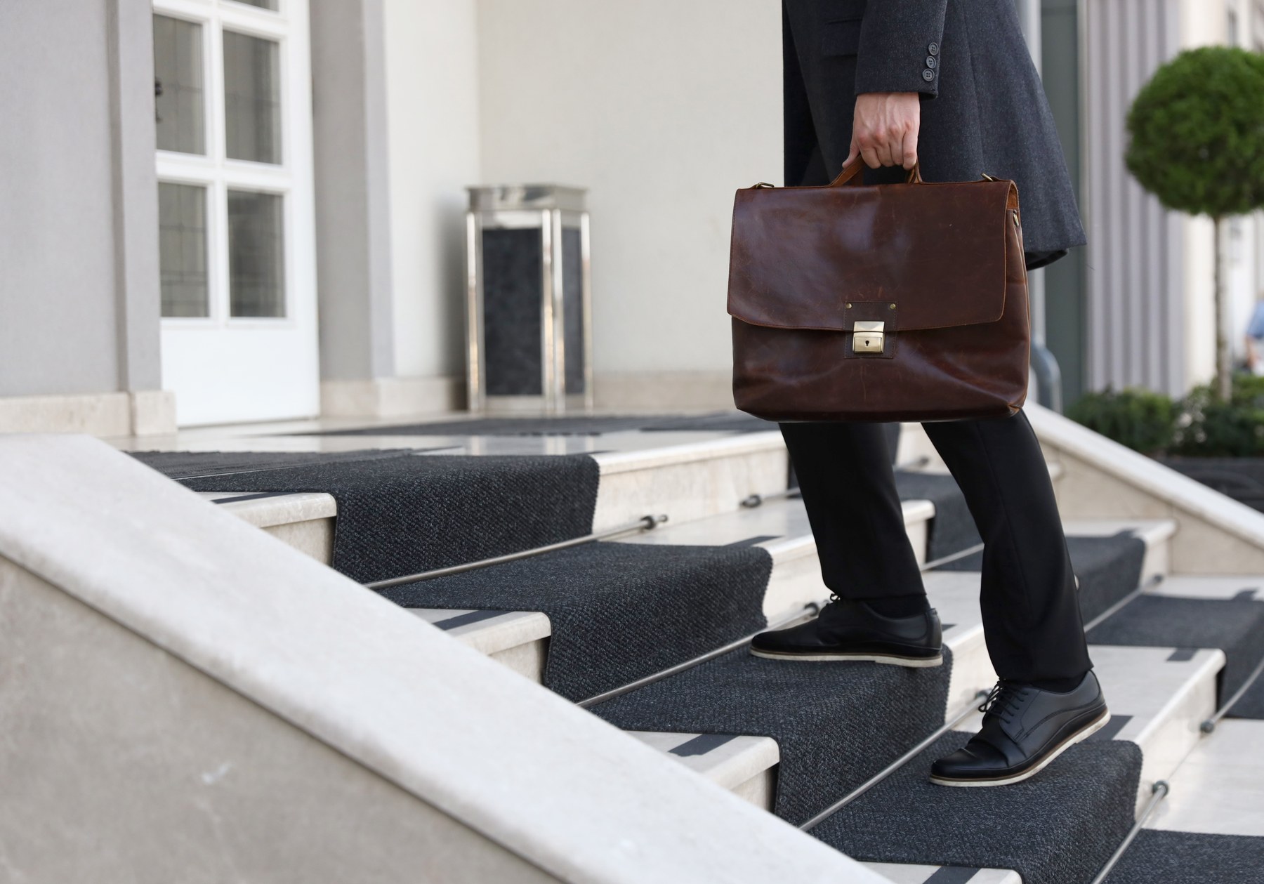 A person in a gray coat climbs carpeted stairs holding a brown leather briefcase. Background shows a white door and decorative plants.