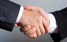 Two people in business suits shaking hands, symbolizing agreement or partnership. Blue-grey background creates a professional tone.