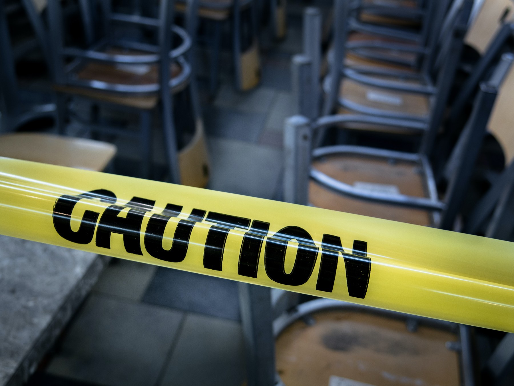 Yellow caution tape in the foreground, with stacked chairs and tables in the background, suggesting a closed or restricted area.